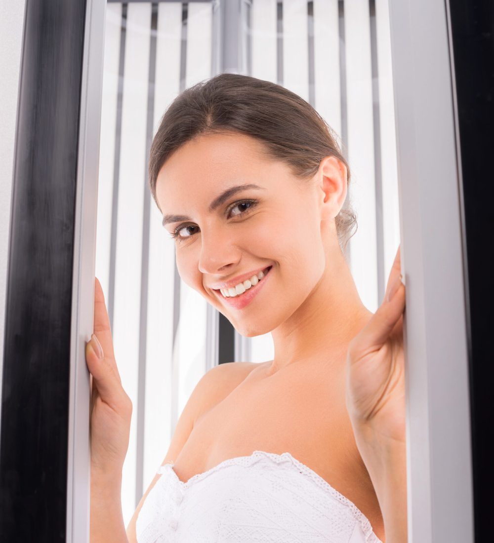 Sunbathing in solarium. Attractive young woman standing in vertical solarium and smiling at camera